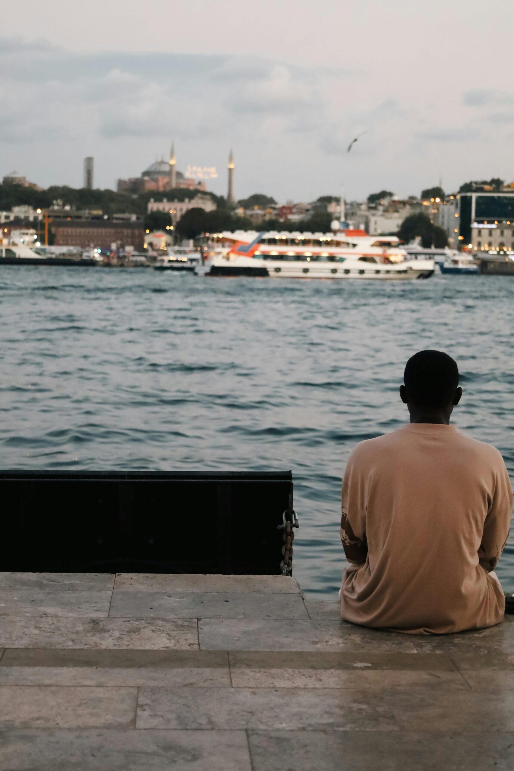 A man sits alone by the water, gazing at Istanbul's iconic skyline.