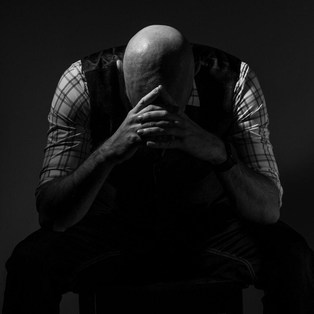 Moody studio portrait of a man in deep thought, hands covering face in monochrome.