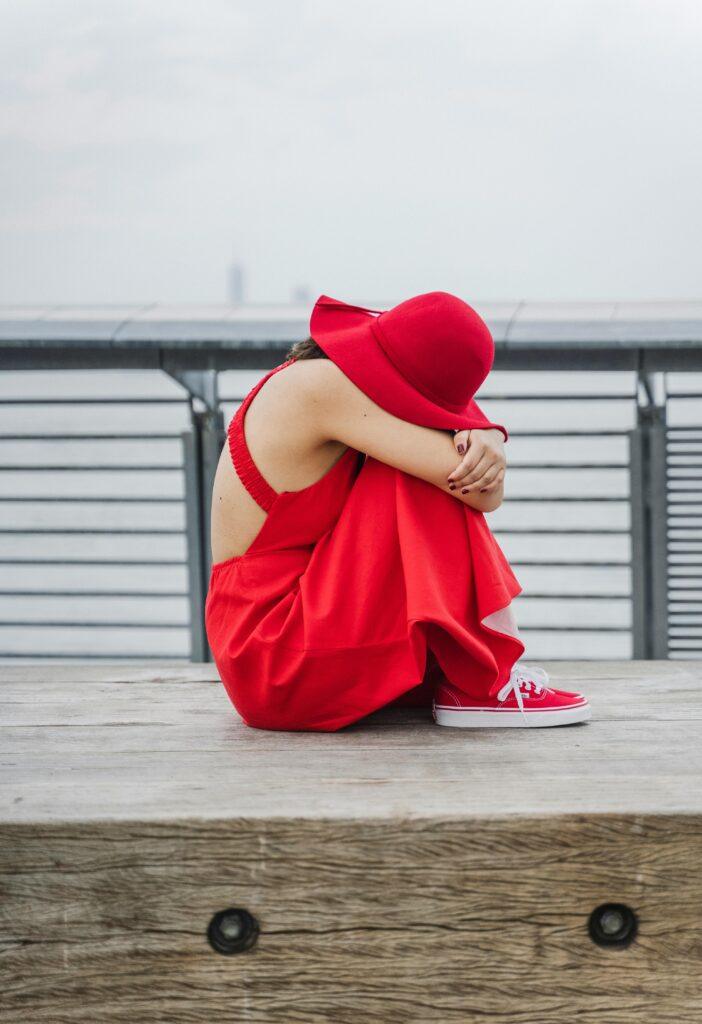 A woman in a vibrant red dress and hat sitting pensively on a bench by the waterfront.